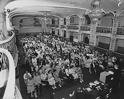 View of the convention general session from a balcony. Attendees are seated alphabetically by state, indicated by signs, as they listen to the speaker at the podium.