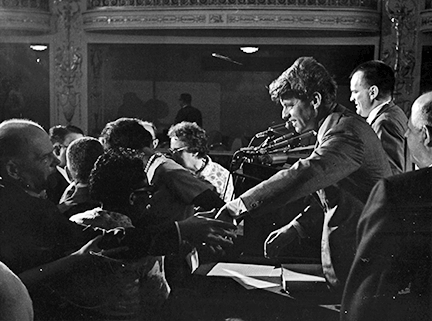 Senator Robert Kennedy seen in profile reaching across a table to shake the hand of a member standing in front of the stage.