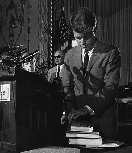 Senator Robert Kennedy examines a stack of three books on the table next to the podium.