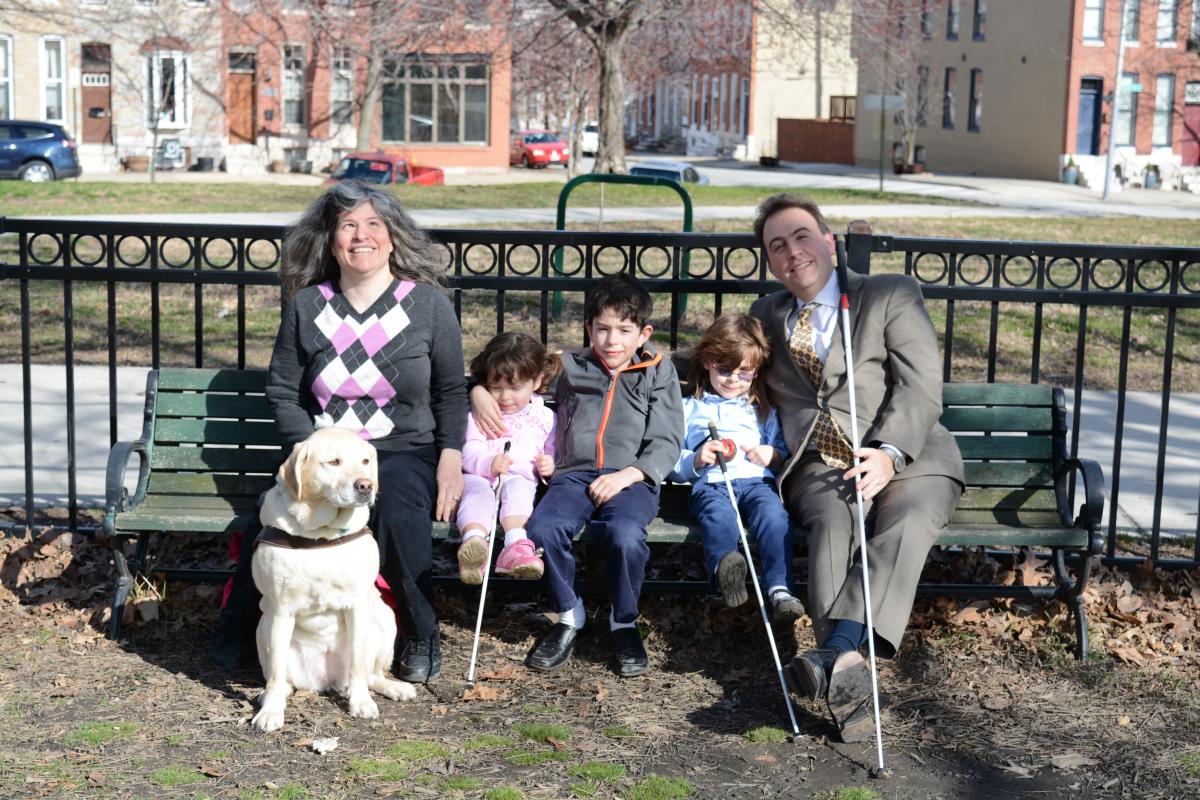 Melissa and Mark Riccobono sit smiling on a park bench with their children.