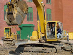 The crane operator crouches in the open door of the crane directing President Maurer as he moves a shovel full of dirt from the hole to a dump truck.