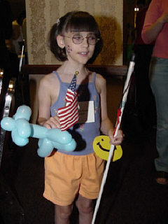 A little girl stands, decorated cane and smiley face in one hand, and a flag and balloon dog in the other.   She has Braille dots on her face.