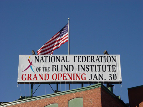 The NFB rooftop sign now reads "National Federation of the Blind Institute Grand Opening January 30th" in large red letters on a white field with a full-color Whozit on the left.   The U.S.flag can be seen flying behind and above the sign.