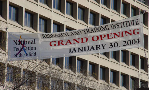 On a banner stretching across the street, the words "National  Federation of the Blind" are flush left and superimposed on Whozit, while on the right are the words "Research and Training Institute Grand Opening January 30, 2004"