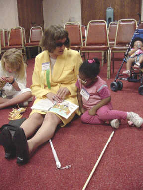Barbara Loos sits  on the floor to read a Braille book to Mikaella Besson.