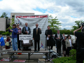 From left an American Sign Language interpreter translates what is being said. Jim Gashel stands with the empty Spellings chair in front of him, listening to Fred Schroeder addressing the crowd, while to the right Joanne Wilson stands at the top of the stairs and NFB President Marc Maurer stands on the steps below her.