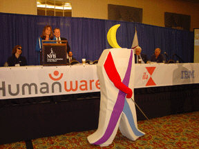 Whozit poses in front of the sponsor banners at the front of the convention hall.