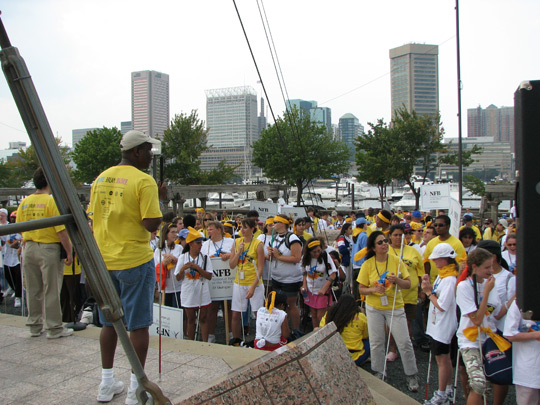 A large crowd of high school students stand listening to a speaker, many wearing yellow Youth Slam T-shirts. We can see lots of state signs being held. The Inner Harbor is visible behind the crowd.