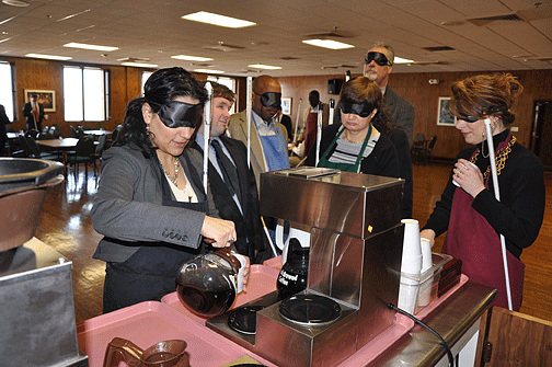 Lourdes Montes-Greenan pours hot coffee while wearing sleepshades. Other Greater Baltimore Committee Leadership Program students wait in line to try their hand at pouring a hot beverage without vision.