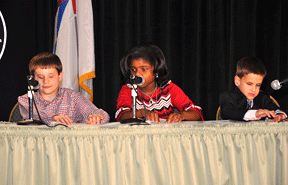 Seated at a table with Braille and microphones in front of them are (left to right) Jason Polansky, Marché Daughtry, and Brandon Pickrel. Marché is reading her part of the resolution.