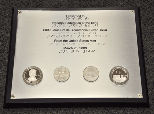The plaque presented to President Maurer by Edmund Moy, director of the United States Mint--the plaque text appears in both print and Braille, and the obverse and the reverse of both the proof (outer) and uncirculated (inner) silver dollars can be seen. The proof coins are mounted in transparent cases for protection, but the uncirculated coins can be touched.