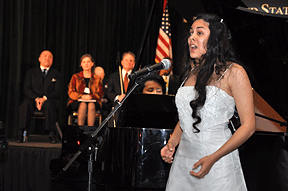 Jessica sings into a floor mic. Visible behind her is her accompanist playing a grand piano, a United States flag on a stand, and dignitaries at the head table.