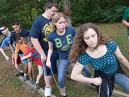 Seven young people stand  in a line, left feet on a two-by-four plank, right feet on a parallel two-by-four  plank, each side controlled independently by ropes held in the students’ hands.  The object is to walk forward by picking up each side in turn, which requires  concentration and much cooperation.