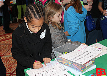 A young girl uses a crayon and a sim-Braille worksheet to write her name while she waits.