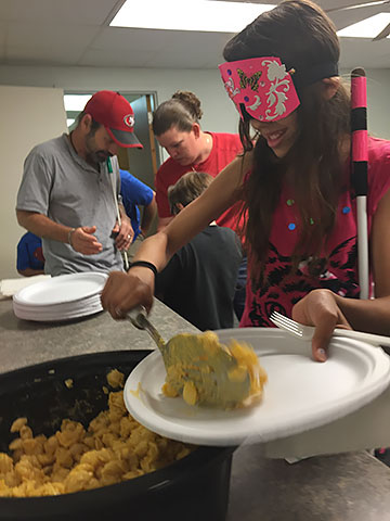 Liz holds her cane and serves herself macaroni and cheese from the buffet line while wearing learning shades.
