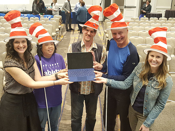 Pictured are Joel Zimba, Jim Gashel and several Microsoft employees all holding a tablet running KNFB Reader. All are wearing Cat in the Hat style hats in observance of Read Across America Day which occurred on March 2.