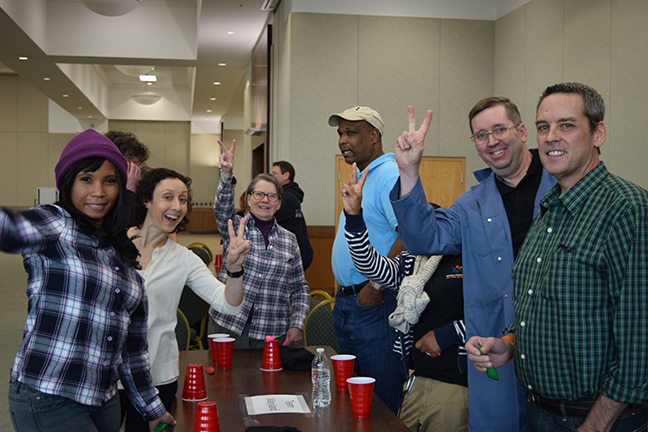 Left to right: Kimie Beverly, Anna Adler, Mary Ellen Jernigan, Anil Lewis, Kathy Douglas (her face is covered by an arm; you can only see her arm up in a peace sign), Bill Jacobs, and Will Schwatka smile together and a few of them hold up the peace sign.