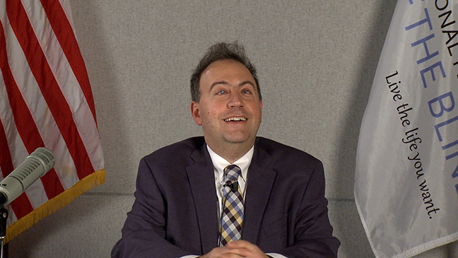 Mark Riccobono smiles while giving the Presidential Release; the American flag and the National Federation of the Blind flag can be seen behind him.