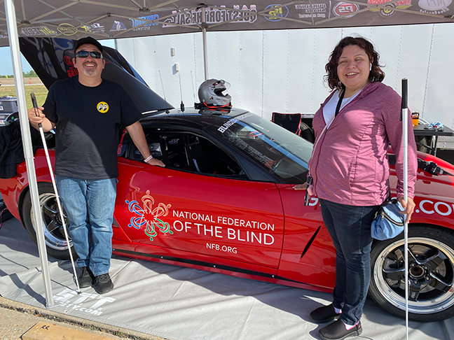 Dan Parker and Danielle McCann, both with white canes, smile next to a red corvette that has the NFB logo on the door.