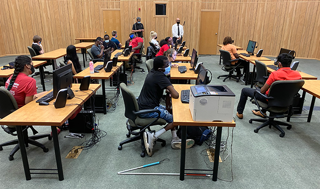 A blind professional speaks to a group of blind students who are seated at computer desks.