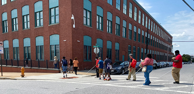A group of blind adults and students cross a street toward the Jernigan Institute, a brick building.