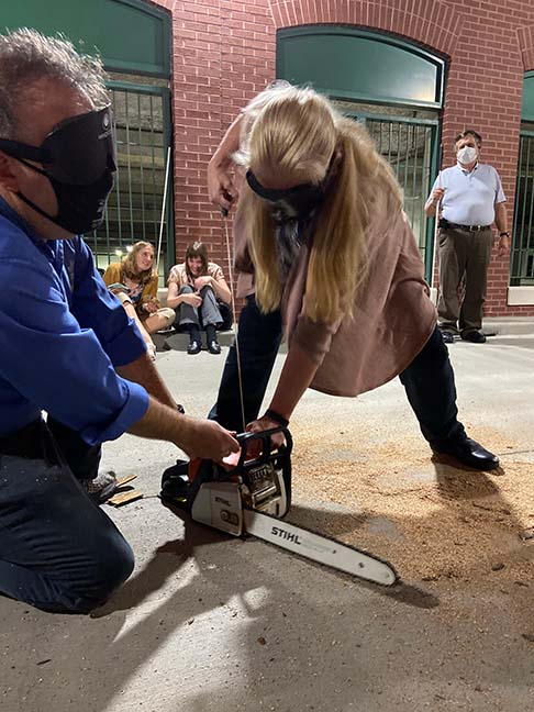 Nikki Wolf bends over and pulls the start cord of the chainsaw. President Riccobono holds the chainsaw steady on the ground for her.