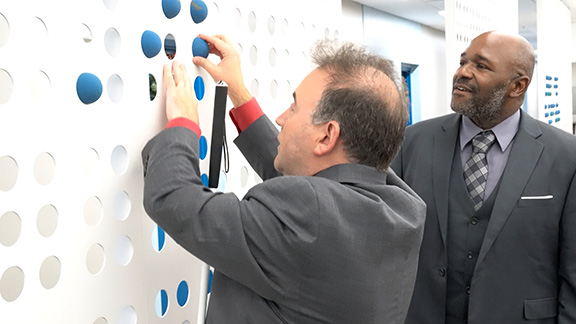 Several tall and wide panels of holes, three down and two across, fill this wall in the NFB Jernigan Institute. Each panel has tennis balls that can be inserted in the holes to create words, sentences, and brief messages. President Riccobono moves some dots to &ldquo;write&rdquo; Braille on the wall for Jason Broughton, the director of the National Library Service for the Blind and Print Disabled.]