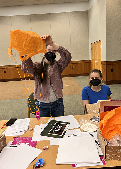 Laura Pitner and Lauren Altman show off the parachute they assembled. One of them holds the parachute up, while the other sits at the table in front of them which is covered with various project materials.