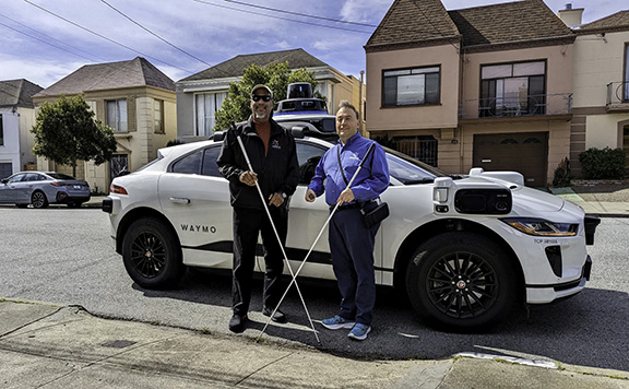 In this photo, Mark and Anil stand smiling in front of the front passenger door of the four-door, white Waymo One vehicle. Their long white canes are crossed and President Riccobono is holding the National Federation of the Blind membership coin.