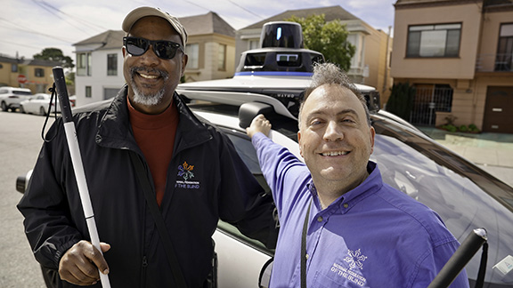 In this close-up photo, Mark and Anil smile joyfully as they lean toward the Waymo One vehicle. The Waymo One is a four-door, white, all-electric Jaguar I-PACE SUV that is equipped with cameras and sensors.