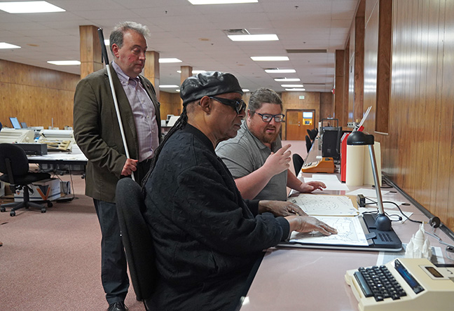 Kennedy Zimnik, a member of the NFB IBTC staff, introduces Stevie Wonder to the Tactonom Reader, which allows you to interact with physical graphics through touch and audio. Mark Riccobono listens in the background.