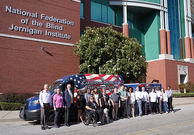 Sandy, Janet, and the National Federation of the Blind staff stand in front of the Caravan, a van equipped with a trailer and decorated with a vibrant design of the American flag featuring other imagery and text. Behind them is a large, four-story, brick building with a sign that reads National Federation of the Blind Jernigan Institute.