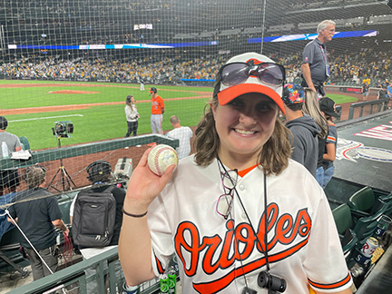 Rose Warner wearing her Orioles gear and holding up a souvenir baseball.