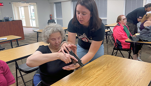 Gun instructor, Haly, stands next to Christine who is seated. Christine is holding a gun, and the instructor is guiding her hands on and around the chamber to get familiar with parts of the gun.