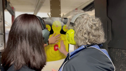 In a booth at the gun range, instructor Haly and Christine touch the yellow target with a red bull&rsquo;s eye that is hanging directly in front of them.