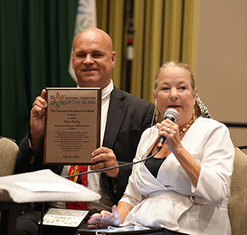 Carla McQuillan speaks about the Distinguished Educator of Blind Students winner Dan Kelley of Ohio. Dan smiles beside her and holds up his award.
