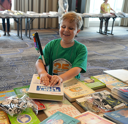 Emerson smiles while reading the cover of a new Braille book. Many other Braille books adorn the tables around him.