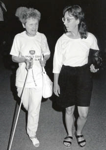 Two adults--one sighted, one blind--walk and converse while the blind adult independently uses her cane.