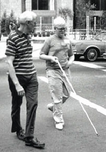 A blind teen uses a cane independently while he and his father cross a street together.