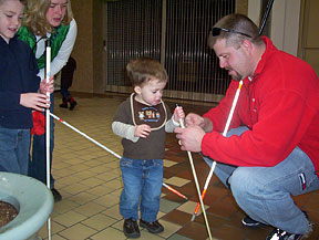 A mother and a boy of about six hold teaching canes as they watch an instructor. The instructor kneels to show a twenty-month-old boy how to hold his tiny cane.