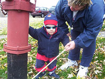 Wearing a look of intense concentration, a little boy explores a fire hydrant with his cane. His mother assists him to tap his cane on the metal.