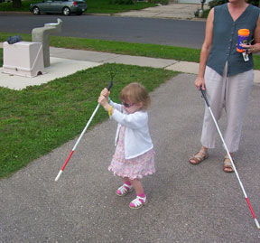 A two-year-old girl experiments with her cane by pounding its tip on the driveway. Her mother, holding her teaching cane, stands back.