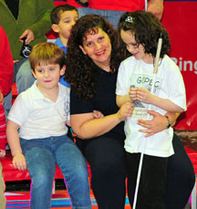Kendra, her mother, and her little brother attend a sports event. Stephanie and R.J. are seated on the bleachers and Kendra stands leaning against her mother's knees.