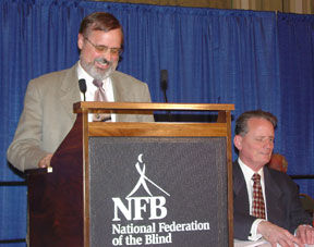 Dr. Matt Maurer stands at a podium while his brother, Dr. Marc Maurer, is seated beside him