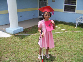 In a pink dress and mortarboard, Torrie stands in front of her house, holding her pre-K diploma