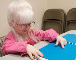 Lyra sits at a table, reading from a Braille book