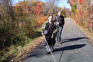 Travel instructor Steve Hastalis walks behind his student as she confidently strides down a paved park trail.