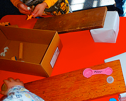 Two children test an inclined plane using objects that include a bottle cap and a measuring spoon.