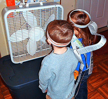 Two small children, wearing hats with paper streamers, stand in front of an electric fan.