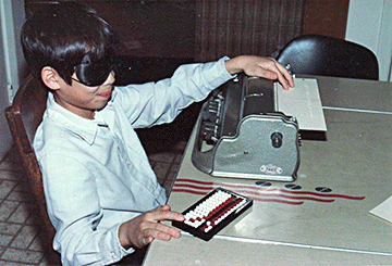 While wearing sleepshades, a boy reads Braille and works out a problem on the abacus.